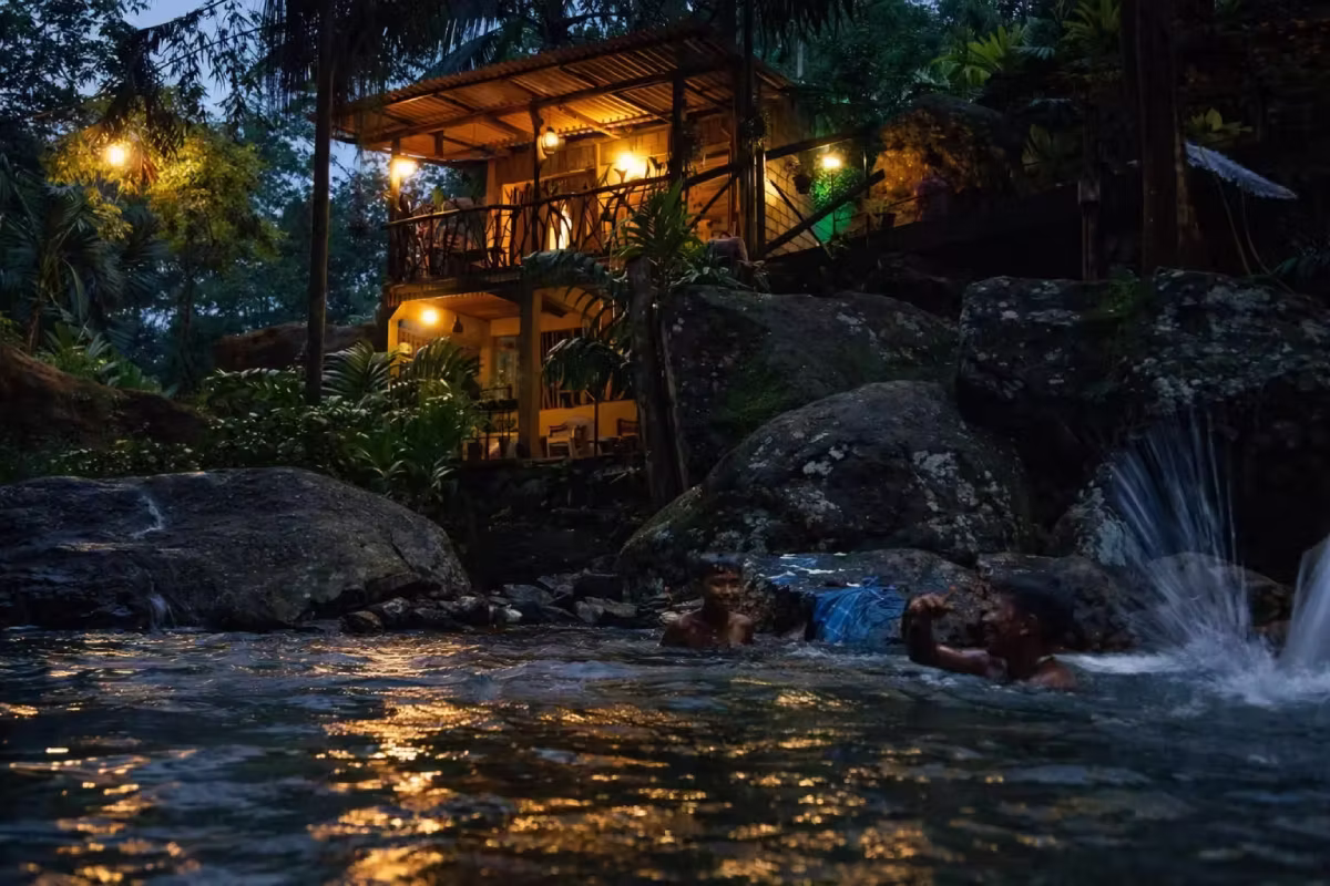 a house illuminated by lights, situated beside a river. Surrounding the house are trees and plants, showcasing a natural setting.